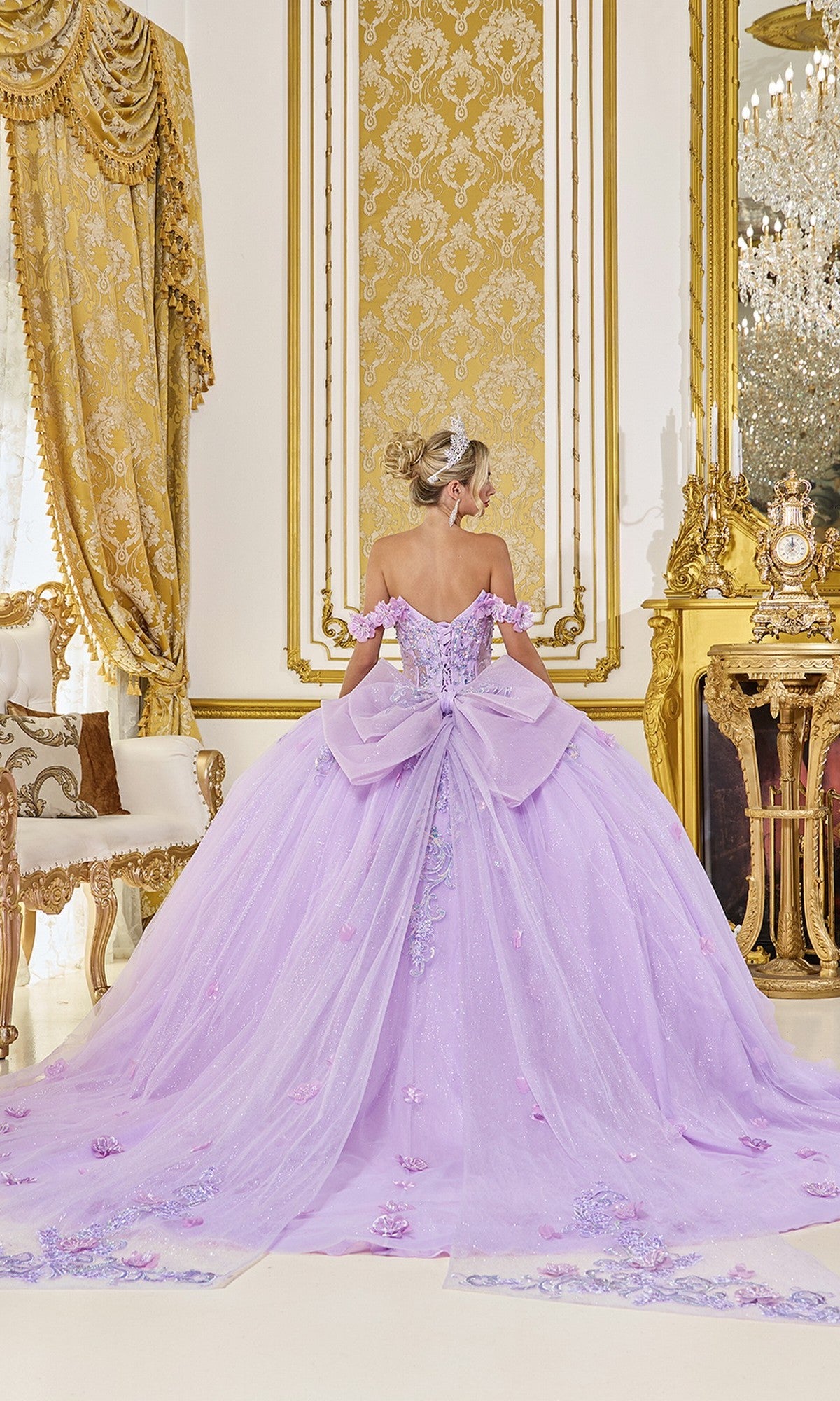 A woman in the Quinceanera Dress 5098 by Dancing Queen, featuring 3D floral accents and a sheer corset bodice, stands in an ornate gold-decorated room with her back to the camera.